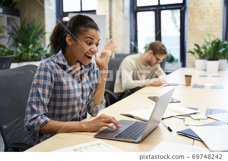 Excited mixed race young woman sitting at the desk in the modern office, looking at laptop screen and celebrating success while her male coworker working on the background. Excited mixed race young woman sitting at the desk in the modern office, looking at laptop screen and celebrating success while her male coworker working on the background. 69748242