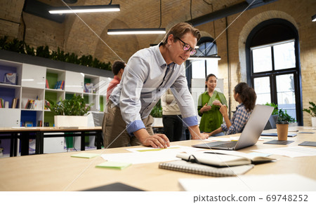 Successful businessman standing in the modern coworking space, looking at laptop screen and thinking about something while his young colleagues talking on the background 69748252
