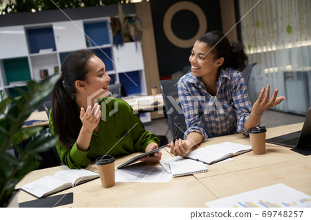 Two young and happy female coworkers sitting at the desk in the modern office and discussing project, working together in coworking space Two young and happy female coworkers sitting at the desk in the modern office and discussing project, working together in coworking space 69748257