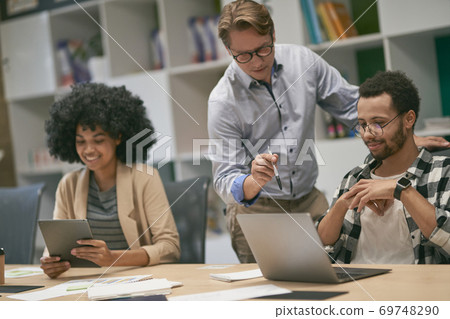 Confident caucasian male boss looking at the laptop screen and discussing project results with two young diverse colleagues sitting at the desk 69748290