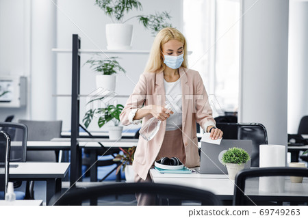 Hygiene and protection of workplace from virus. Attractive millennial woman in protective mask disinfects laptop in office interior 69749263