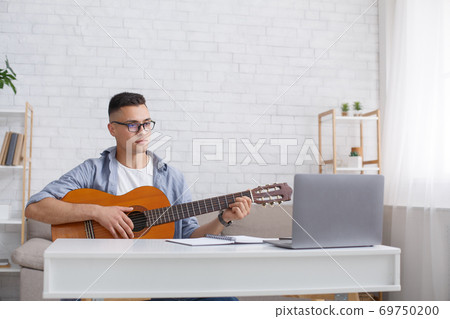 Modern technology and app for learning music in free time at home. Concentrated guy in glasses plays the guitar and looks at laptop in interior of living room 69750200