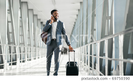 Cheerful Black Businessman Chatting On Phone Walking In Airport, Panorama Cheerful Black Businessman Chatting On Phone Walking In Airport, Panorama 69750390