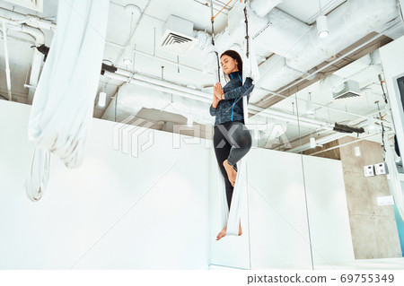 Ariel yoga. Young caucasian woman in sportswear practicing fly yoga in studio, standing on the one leg in white hammock and meditating 69755349