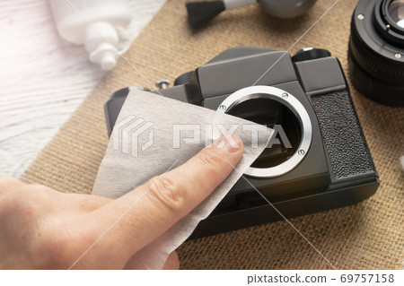 The hand cleans the camera with a napkin from dust and dirt on a sack and a wooden table. a blower with a brush and a special fluid are nearby. Maintenance of photographic equipment at home The hand cleans the camera with a napkin from dust and dirt on a sack and a wooden table. a blower with a brush and a special fluid are nearby. Maintenance of photographic equipment at home 69757158
