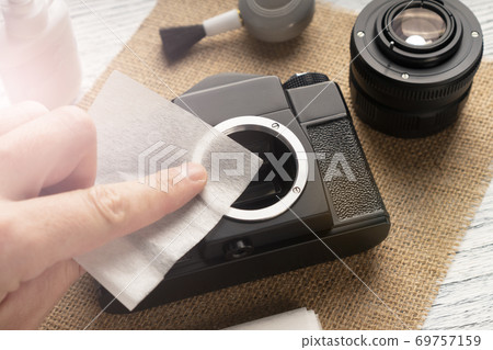 The hand cleans the camera with a napkin from dust and dirt on a sack and a wooden table. a blower with a brush and a special fluid are nearby. Maintenance of photographic equipment at home The hand cleans the camera with a napkin from dust and dirt on a sack and a wooden table. a blower with a brush and a special fluid are nearby. Maintenance of photographic equipment at home 69757159
