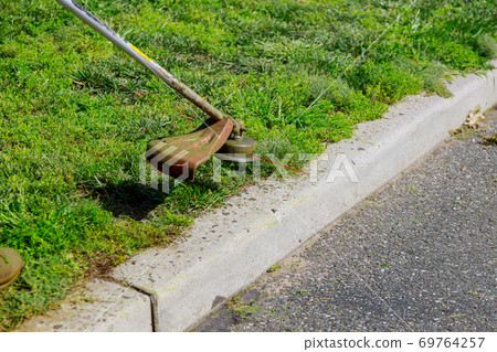 A lawn mower mows fresh, green grass on the lawn near, a municipal worker  69764257