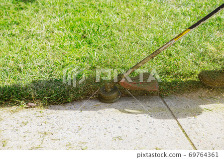 A lawn mower mows fresh, green grass on the lawn near, a municipal worker with lawn mower in his hand. 69764361