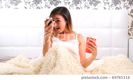 Attractive young woman gets up in bed with a cup of coffee and an alarm clock. The woman overslept Attractive young woman gets up in bed with a cup of coffee and an alarm clock. The woman overslept 69765318