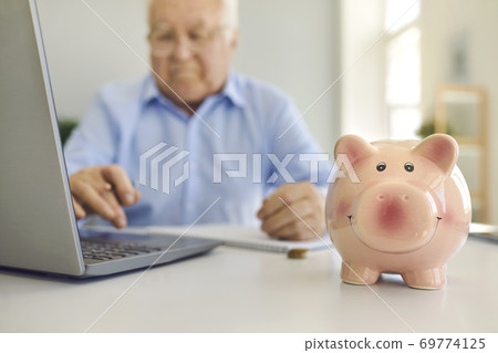 Piggy bank standing on desk with blurred senior man using laptop to pay bills online in background 69774125