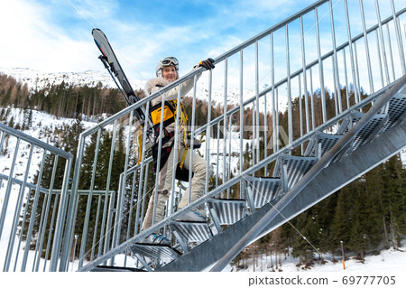Portrait of young adult female brave skier tourist with skiing equipment enjoy having fun going to ride extreme suspended zipline wire flying over gorge canyon in mountains on background. Adrenaline Portrait of young adult female brave skier tourist with skiing equipment enjoy having fun going to ride extreme suspended zipline wire flying over gorge canyon in mountains on background. Adrenaline 69777705