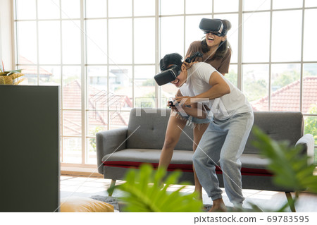 Young happy Asian couple playing video games wearing virtual reality glasses in their apartment Young happy Asian couple playing video games wearing virtual reality glasses in their apartment 69783595