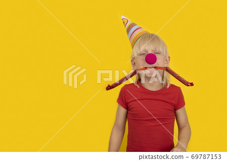 Boy blowing in festive whistle. Portrait of child with clown nose and party hat. Boy blowing in festive whistle. Portrait of child with clown nose and party hat. 69787153