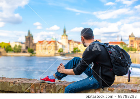 A tourist guy with a backpack sits on the embankment in Prague overlooking the Vltava river 69788288
