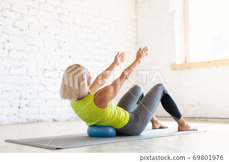 Adult caucasian woman practice pilates indoor. Seated rolls up with small fit ball on a mat, abs and back drill, in loft white studio, selective focus. 69801976