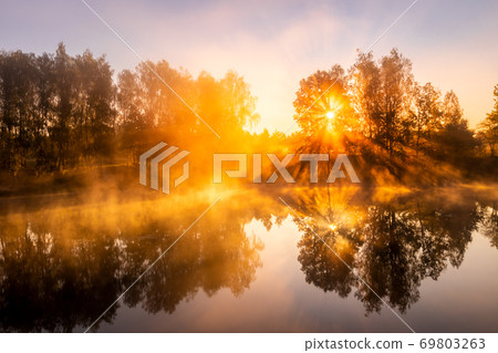 Golden misty sunrise on the pond in the autumn morning. Trees with rays of the sun cutting through it, reflected in the water. Golden misty sunrise on the pond in the autumn morning. Trees with rays of the sun cutting through it, reflected in the water. 69803263
