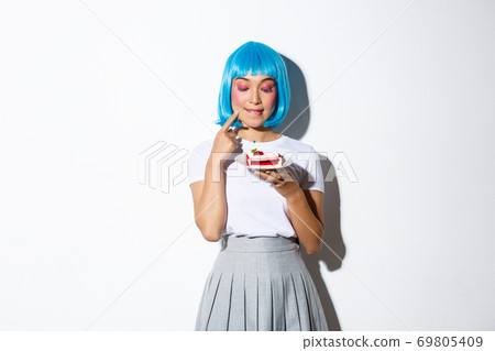 Image of coquettish fashion girl in blue wig looking with temptation at cake, standing over white background 69805409