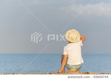 Child in a straw hat sitting on beach and looking at the distance. Holidays. Back view Child in a straw hat sitting on beach and looking at the distance. Holidays. Back view 69809744
