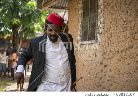 An African Older Man in Red Muslim Taqiyyah Fez Hat And Blazer on white Dress Moving a stick for lame people Near the Basic Hut with Thatched roof in Small Remote Village in Tanzania, Pemba island An African Older Man in Red Muslim Taqiyyah Fez Hat And Blazer on white Dress Moving a stick for lame people Near the Basic Hut with Thatched roof in Small Remote Village in Tanzania, Pemba island 69818107