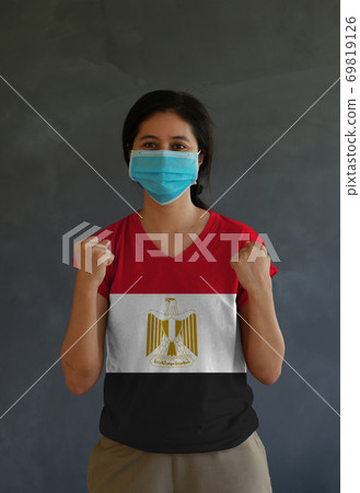 Woman wearing hygienic mask and wearing Egypt flag colored shirt and standing with raised both fist Woman wearing hygienic mask and wearing Egypt flag colored shirt and standing with raised both fist 69819126