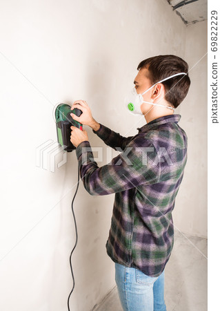 Man in protective mask working with electric sander to smooth plaster wall surface, room renovation concept Man in protective mask working with electric sander to smooth plaster wall surface, room renovation concept 69822229