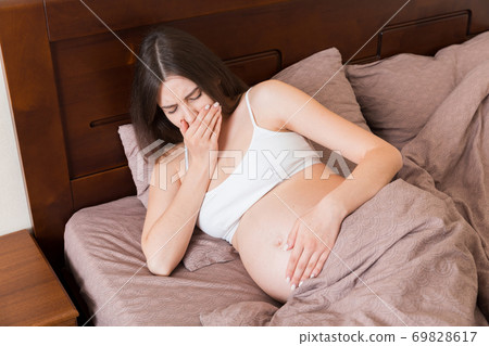 Pregnant suffering with nausea. Pregnancy symptoms, expectation toxicosis. Young vomiting woman sitting on bed. parenthood concept Pregnant suffering with nausea. Pregnancy symptoms, expectation toxicosis. Young vomiting woman sitting on bed. parenthood concept 69828617
