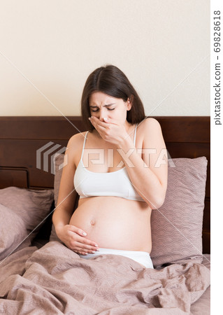 Pregnant suffering with nausea. Pregnancy symptoms, expectation toxicosis. Young vomiting woman sitting on bed. parenthood concept Pregnant suffering with nausea. Pregnancy symptoms, expectation toxicosis. Young vomiting woman sitting on bed. parenthood concept 69828618