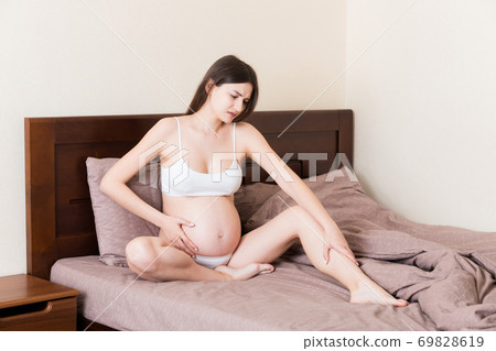 Pretty pregnant woman hands massaging leg sitting on bed Pretty pregnant woman hands massaging leg sitting on bed 69828619