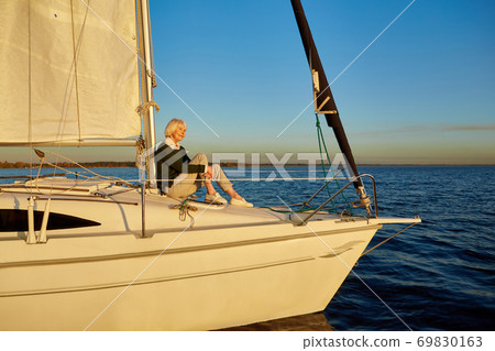 Enjoying luxury life. Beautiful senior woman sitting on the side of sailboat or yacht deck floating in the calm blue sea at sunset, looking away and smiling Enjoying luxury life. Beautiful senior woman sitting on the side of sailboat or yacht deck floating in the calm blue sea at sunset, looking away and smiling 69830163