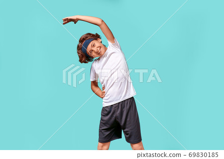 Warming up. Happy teenage boy in sportswear looking away and smiling while stretching his body before workout, standing isolated over blue background 69830185
