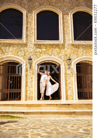 couple dressed in white, stands in arch of house. 69832277