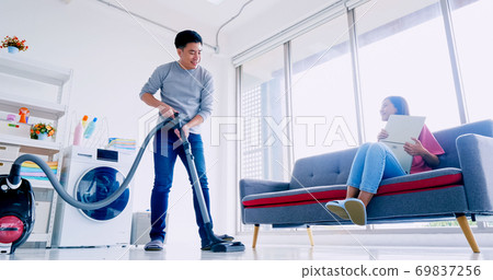 Husband cleaning floor with vacuum cleaner while wife working with laptop on sofa. Husband cleaning floor with vacuum cleaner while wife working with laptop on sofa. 69837256