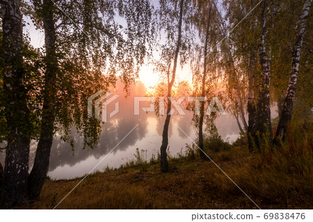 Golden misty sunrise on the pond in the autumn morning. Trees with rays of the sun cutting through it, reflected in the water. Golden misty sunrise on the pond in the autumn morning. Trees with rays of the sun cutting through it, reflected in the water. 69838476