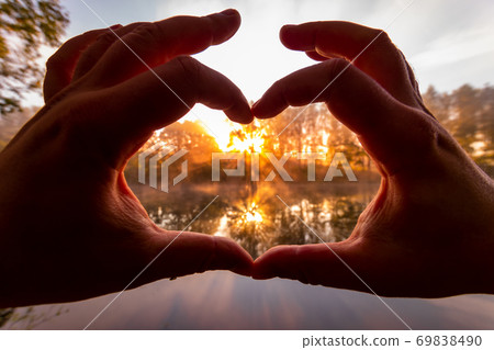 Male hands making a heart-shaped gesture against the background of sunset or sunrise on a river or lake. Male hands making a heart-shaped gesture against the background of sunset or sunrise on a river or lake. 69838490