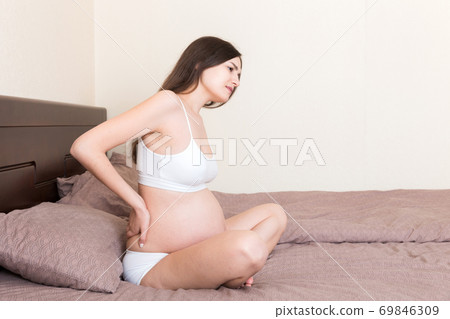 beautiful pregnant woman touching her tummy and keeping one hand on her back at home on bed beautiful pregnant woman touching her tummy and keeping one hand on her back at home on bed 69846309