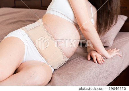 closeup pregnant woman dresses bandage on belly at home on bed. Orthopedic abdominal support belt closeup pregnant woman dresses bandage on belly at home on bed. Orthopedic abdominal support belt 69846350