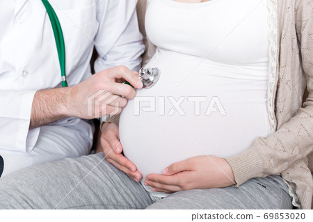 Doctor is checking pregnant woman's belly with a stethoscope. Physician is listening to a heartbeat of a baby Doctor is checking pregnant woman's belly with a stethoscope. Physician is listening to a heartbeat of a baby 69853020