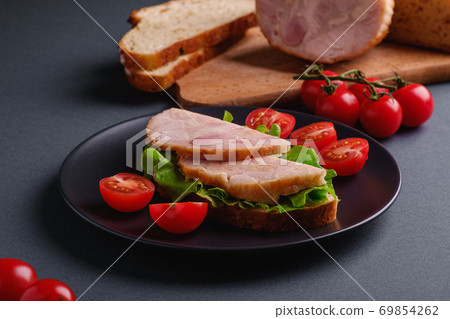 Sandwich with turkey ham meat, green salad and fresh cherry tomatoes slices on black plate near to ingredients on cutting board, grey minimal background, angle view macro 69854262