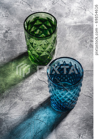 Two geometric glass cup with water in blue and green colors with colorful shadow light rays on stone concrete background, angle view Two geometric glass cup with water in blue and green colors with colorful shadow light rays on stone concrete background, angle view 69854616