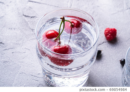 Fresh cold sparkling water drink with cherry, raspberry and currant berries in transparent glass on stone concrete background, summer diet beverage, angle view macro Fresh cold sparkling water drink with cherry, raspberry and currant berries in transparent glass on stone concrete background, summer diet beverage, angle view macro 69854984