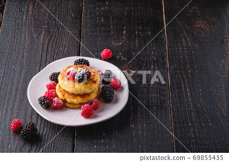 Cottage cheese pancakes and powdered sugar, curd fritters dessert with raspberry and blackberry berries in plate on dark black wooden background, angle view copy space Cottage cheese pancakes and powdered sugar, curd fritters dessert with raspberry and blackberry berries in plate on dark black wooden background, angle view copy space 69855435