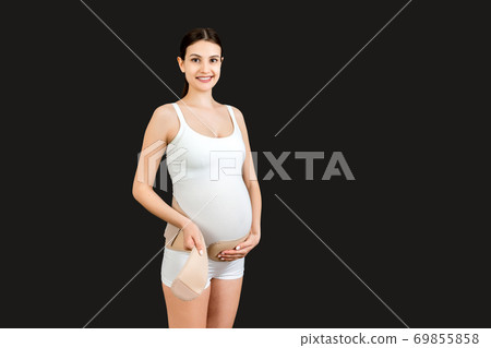 Portrait of pregnant woman putting on pregnancy bandage at black background with copy space. Orthopedic abdominal support belt concept Portrait of pregnant woman putting on pregnancy bandage at black background with copy space. Orthopedic abdominal support belt concept 69855858