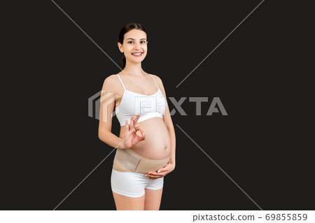 Portrait of pregnant woman in underwear on the third trimester wearing pregnancy belt and showing okay sign at black background. Orthopedic abdominal support belt concept with copy space 69855859
