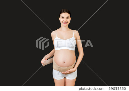 Portrait of pregnant woman in underwear dressing pregnancy corset on the third trimester at black background with copy space. Orthopedic abdominal support belt concept Portrait of pregnant woman in underwear dressing pregnancy corset on the third trimester at black background with copy space. Orthopedic abdominal support belt concept 69855860