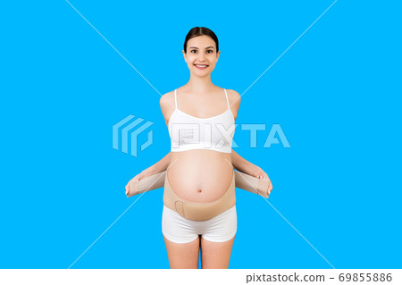 Portrait of pregnant woman in underwear putting on supporting bandage for reducing backache at blue background with copy space. Orthopedic abdominal support belt concept Portrait of pregnant woman in underwear putting on supporting bandage for reducing backache at blue background with copy space. Orthopedic abdominal support belt concept 69855886