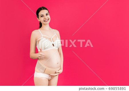 Portrait of young pregnant woman in underwear wearing bandage on her belly and showing thumb up sign at pink background with copy space. Orthopedic abdominal support belt concept 69855891