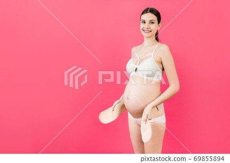 Portrait of pregnant woman in underwear dressing pregnancy corset on the third trimester at pink background with copy space. Orthopedic abdominal support belt concept Portrait of pregnant woman in underwear dressing pregnancy corset on the third trimester at pink background with copy space. Orthopedic abdominal support belt concept 69855894