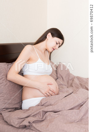 Young pretty pregnant woman, in pain, sitting on bed in bedroom, preparing to give a birth Young pretty pregnant woman, in pain, sitting on bed in bedroom, preparing to give a birth 69870412