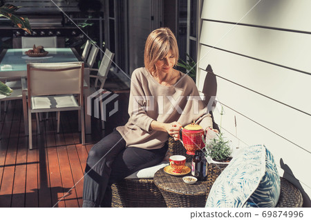 Middle-aged woman pouring tea from teapot to cup on the balcony Middle-aged woman pouring tea from teapot to cup on the balcony 69874996