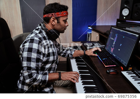 Young man, male artist looking focused while playing keyboard synthesizer, sitting in recording studio Young man, male artist looking focused while playing keyboard synthesizer, sitting in recording studio 69875356